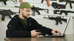 Man in green hat sits at desk with laptop to update pistol inventory.