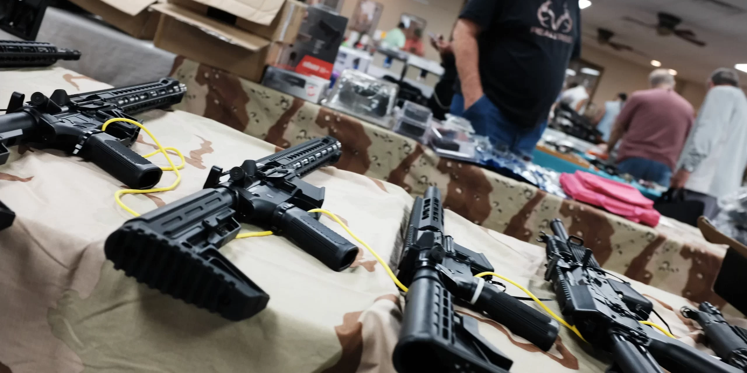 A busy gun show booth with vendors showcasing firearms and gear, while customers browse and transact using mobile sales technology.