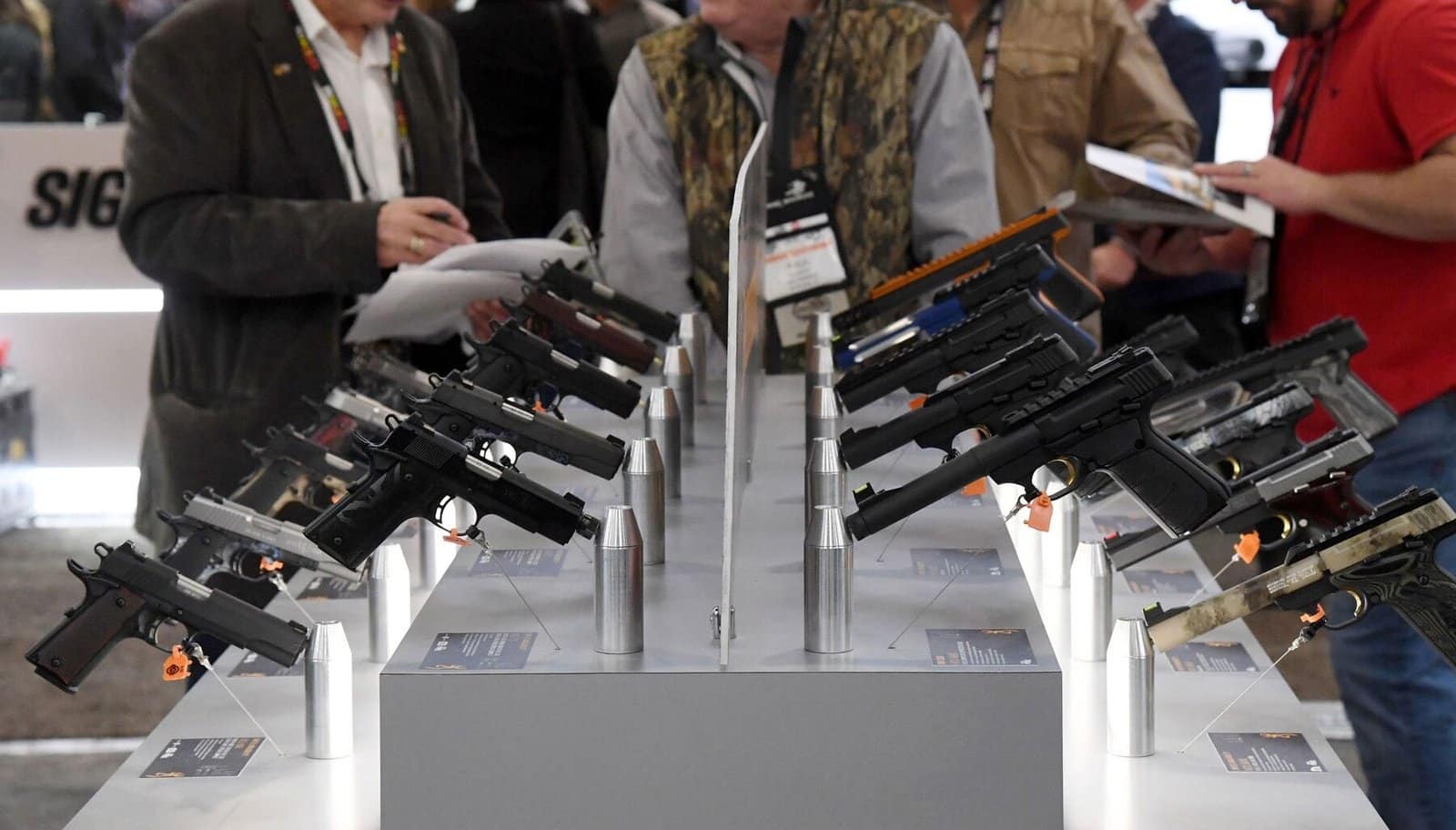 Handguns neatly displayed on a white stand at a Gun Show, with attendees in the background examining firearms—RapidGunSystems.com gun store consignment software.
