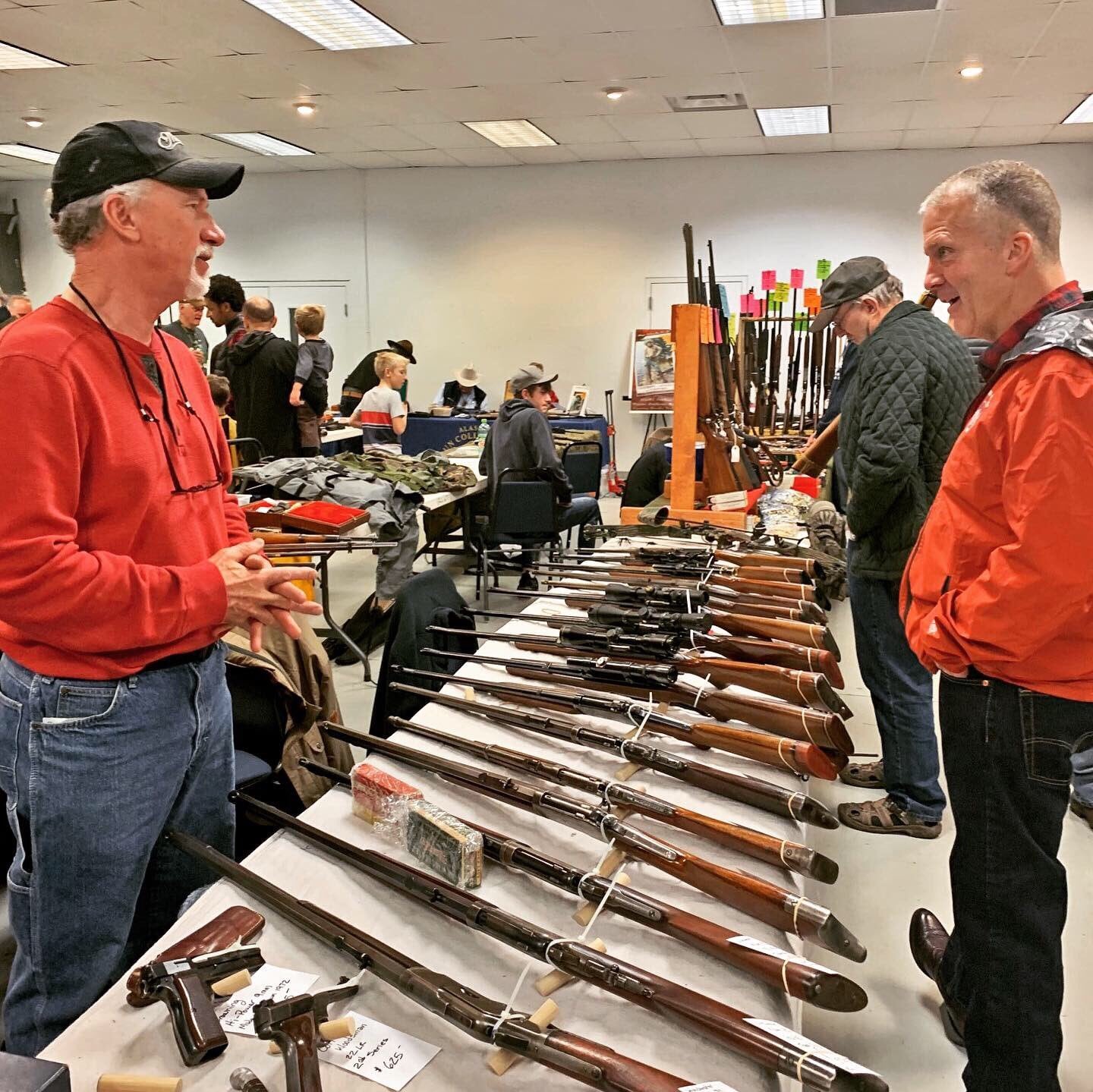 Two men discuss firearms at a gun show booth, surrounded by rifles and handguns, while attendees browse RapidGunSystems event.