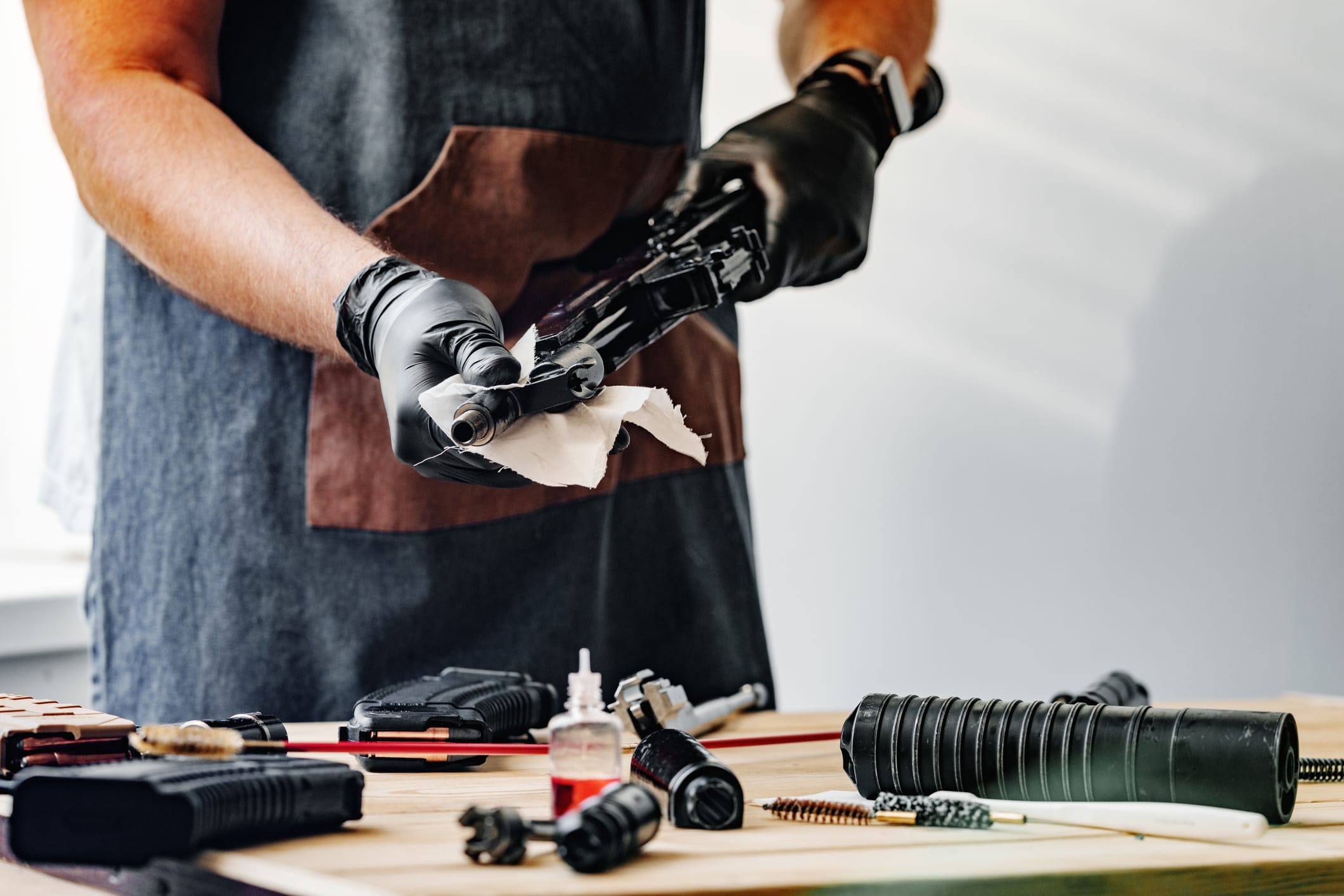 A gunsmith in black gloves and apron cleans a rifle on a workbench with parts, tools, cleaning supplies, and gunsmithing software to easily manage open workorders.