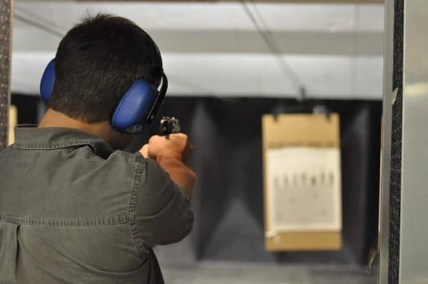 At an indoor range, a shooter in blue earmuffs fires at a target, using Rapid Gun Systems Shooting Range Management Software for efficient range management.