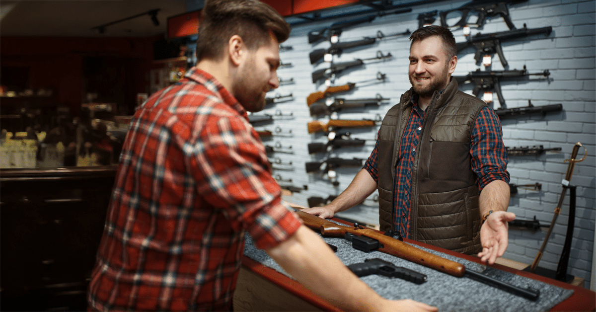 Inside a gun store, two men discuss shooting range management software at a counter displaying firearms and rifles working with the advanced Gun Store POS Features with Offline Sales for Gun Stores