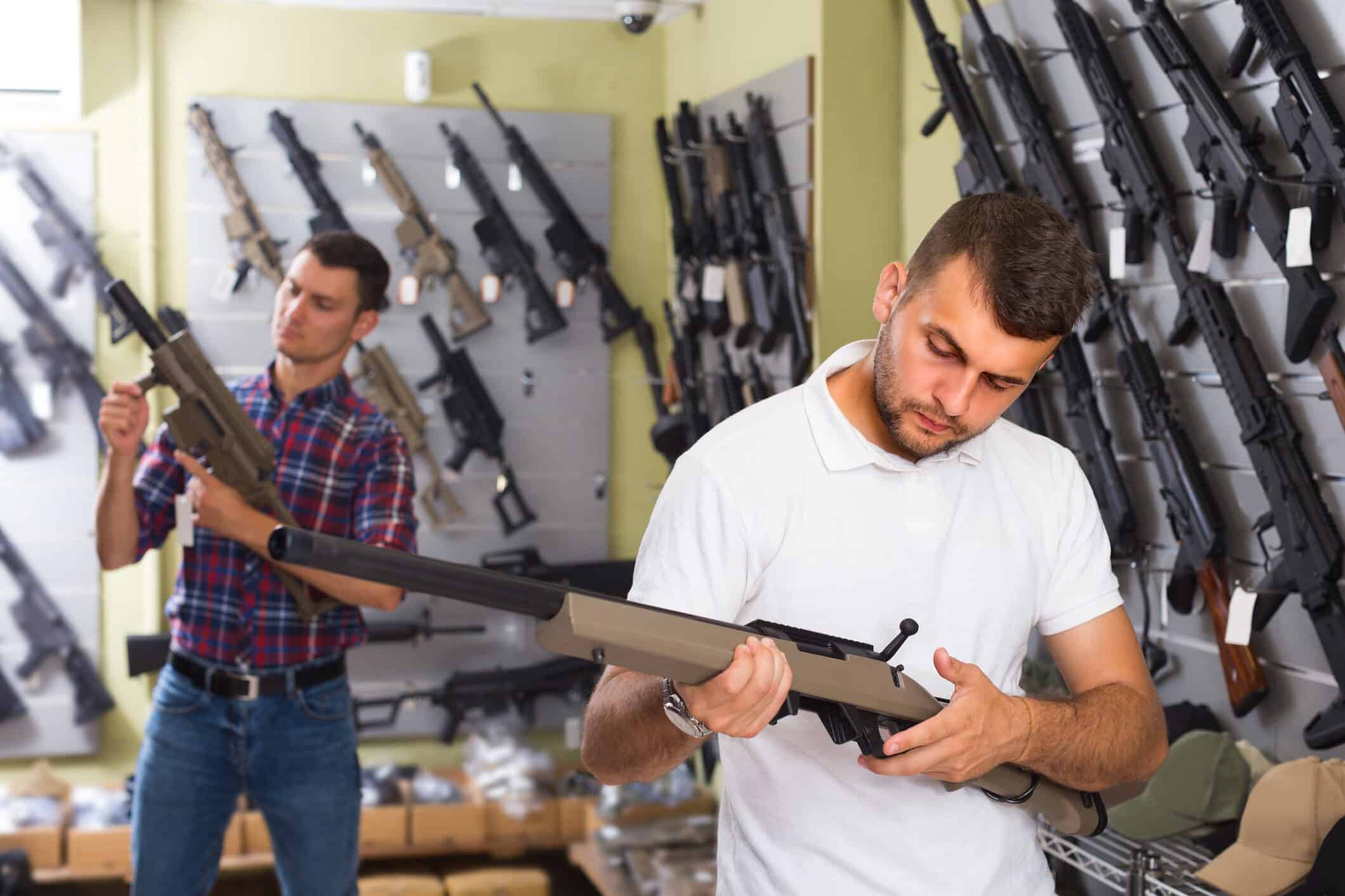Inside a gun store, two men inspect rifles; firearms line the walls and signs promote Loyalty Programs for Gun Stores managed within the rapid gun systems pos